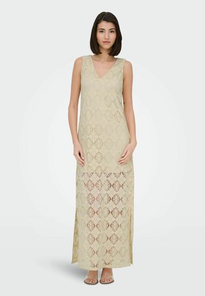 Woman wearing a sleeveless beige lace maxi dress with a V-neck, standing with hands gently touching, on a plain light background.