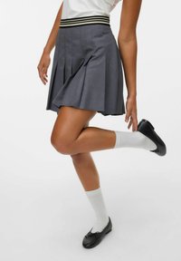 Personne portant une jupe plissée grise, des chaussettes blanches, des ballerines noires, levant une jambe avec la main près du pied, sur fond blanc.