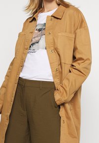 Brun knappad skjorta med bröstfick över en vit grafisk t-shirt, ihop med olivgröna byxor. Slät tyg med avslappnad passform.