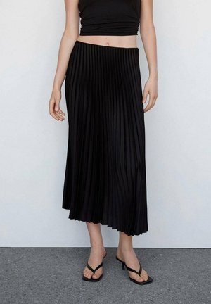 Jupe midi plissée noire avec une silhouette fluide, fabriquée en tissu lisse. Portée avec un haut noir ajusté et des tongs noires plates.