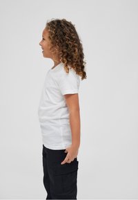Vaikas šonu nufotografuotas, vilkintis baltą trumpą rankovę T-shirt ir juodas cargo kelnes. Matomi garbanoti plaukai, kūnas šiek tiek pasukęs.