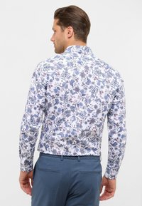 Camicia a maniche lunghe floreale bianca con motivi blu e viola, dotata di colletto classico e chiusura con bottoni. Indossata con pantaloni azzurri chiari.