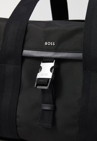 Fekete nylon sporttáska állítható pántokkal, fém ezüst csattal és márkázott logóval. Simább tapintás, minimalista dizájn.