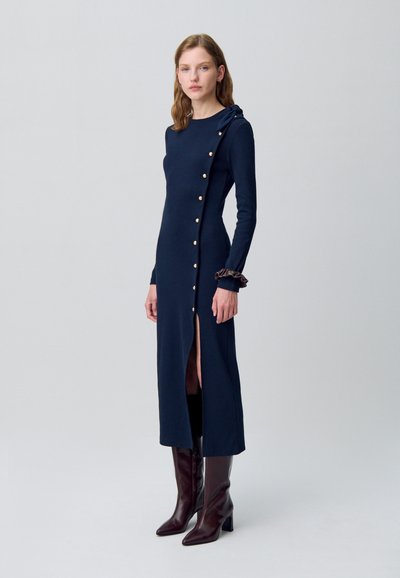 Robe longue marine à manches longues avec une fente sur le côté, des boutons dorés sur le devant et un accent texturé sur la manche. Associée à des bottes à talons marron.