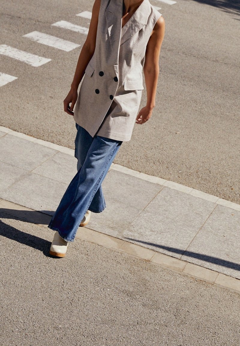 Personne marchant dans la rue portant un blazer beige sans manches, un jean bleu à jambes larges et des chaussures blanches près d'un passage pour piétons.