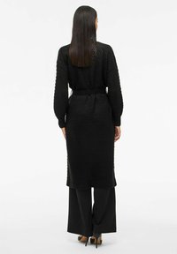 Cardigan long noir en tricot avec un motif texturé et des poignets côtelés, associé à un pantalon large noir et des talons hauts.