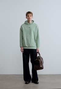 Φωτεινό πράσινο φούτερ με κουκούλα και τσέπη καγκουρό, συνοδευόμενο από μαύρο παντελόνι με φαρδιά μπατζάκια και καφέ δερμάτινη τσάντα με στρογγυλή λαβή.