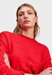 Pull rouge avec un col rond, présentant de petites embellissements brillants. Texture douce avec une coupe décontractée. Boucles d'oreilles en forme de créole dorées portées.