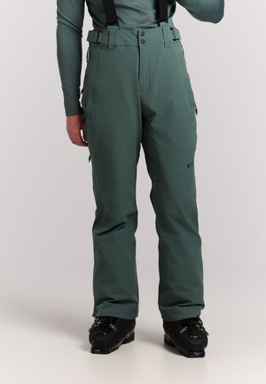 Mann trägt grüne wasserdichte Skihose mit schwarzen Hosenträgern und schwarzen Skischuhen, steht vor weißem Hintergrund.