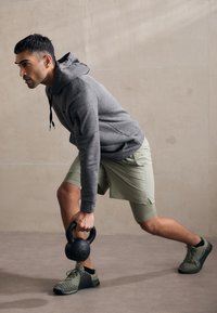 Pilka hoodie su raišteliais, šviesiai žali šortai ir juodas kettlebellas sportinėje pozoje, prieš neutralaus tonų foną.