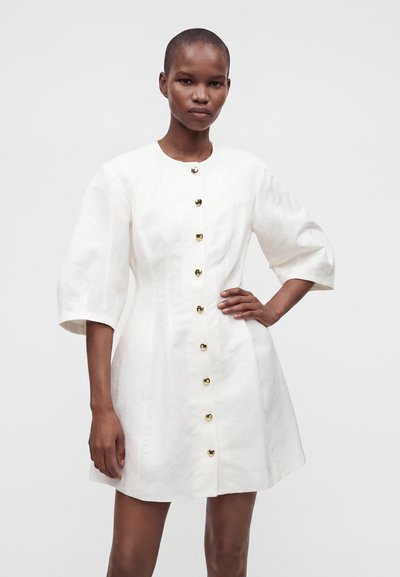 Femme portant une robe blanche avec des manches trois-quarts bouffantes et des boutons dorés sur le devant, debout avec une main sur la hanche.