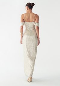 Dolga, oprijeta obleka iz srebrnih bleščic, z odprtimi rameni in tankimi naramnicami. Tkanina ima gladko, odsevno teksturo.