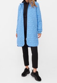 Ljusblå quiltad lång rock med huva, med dragkedjestängning och sidofickor. Buren över en svart hoodie och kombinerad med svarta byxor och sneakers.