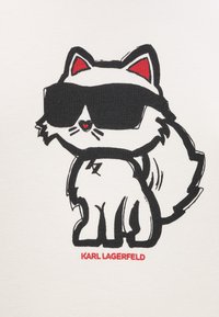 Biały materiał z kocurem w stylu kreskówkowym, czarnym konturem, czerwonymi akcentami na uszach i dużymi czarnymi okularami przeciwsłonecznymi, z napisem "KARL LAGERFELD" poniżej.