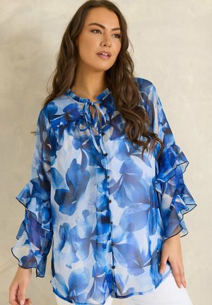 Vrouw met lang bruin haar, gekleed in een doorschijnend, blauw bloemenblouse met knopen en geruite mouwen, staand tegen een neutrale achtergrond.