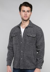 Grijze shirtjas met visgraatstructuur, voorzien van twee borstzakken, knoopsluiting en lange mouwen. De stof lijkt dik en warm te zijn.