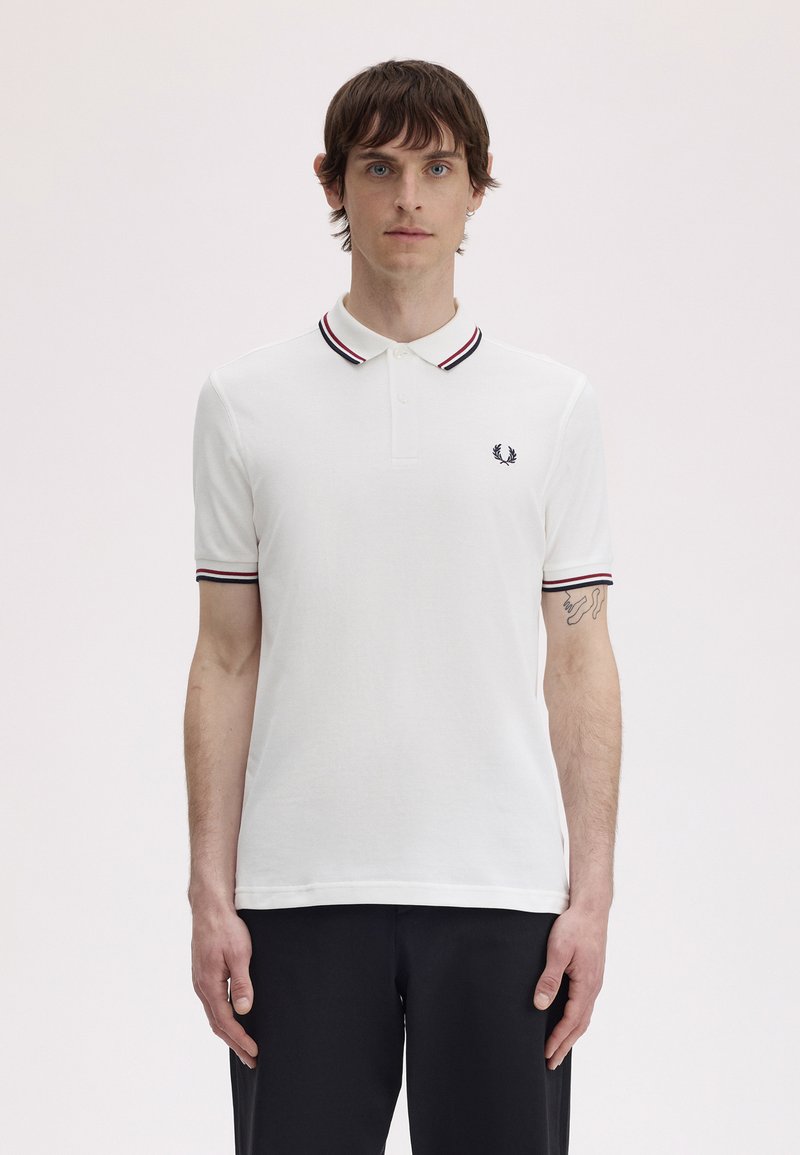 Hombre con camisa polo blanca con cuello y mangas a rayas negras y rojas, de pie contra un fondo claro y liso.