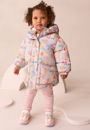 Kleinkind mit lockigem Haar steht und trägt eine bunte, mit Tiermotiven bedruckte Steppjacke, rosa Hose und pastellfarbene Sneakers vor neutralem Hintergrund.