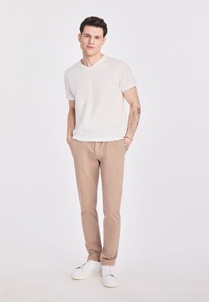 Biała koszulka v-neck, beżowe spodnie i białe sneakersy. Minimalistyczny design, miękki materiał, dopasowany krój. Tatuaż widoczny na lewym przedramieniu.