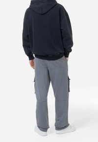 Man i svart hoodies, grå cargobyxor med sidofickor och vita sneakers står med händerna i fickorna och tittar bort.