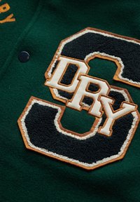 Groene viltstof met een geborduurd "S" en "DRY" in crème en zwart, omrand door bruine accenten. Knopsluiting zichtbaar.