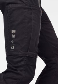 Černé pracovní kalhoty s hrubou texturou, vybavené detaily kapes, bílým prošíváním a potištěnými čísly na stehně. Zip na boku.