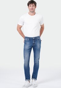 Homme se tenant debout avec les mains derrière le dos, portant un t-shirt blanc, un jean bleu retroussé et des baskets blanches sur un fond clair.