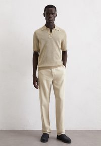 Beige knitted polo shirt with short sleeves, paired with cream-colored trousers and dark loafers, set against a neutral backdrop.