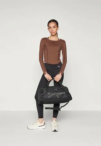 Mujer con una camiseta de manga larga marrón y leggings negros sosteniendo una bolsa de deporte negra, vestida con zapatillas blancas, de pie contra un fondo claro y liso.