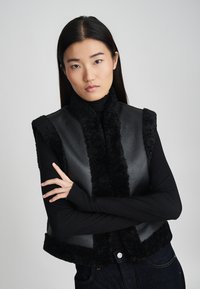 Μαύρο δερμάτινο αμάνικο γιλέκο με υφή από μαύρη γούνα στον λαιμό και στις άκρες, με άνετη εφαρμογή και διακριτική λάμψη. Φορεμένο πάνω από μια μαύρη μπλούζα.