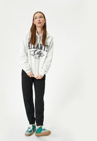 Witte sweatshirt met zwarte grafische tekst, gecombineerd met zwarte broeken. Schoenen zijn teal en grijs, met witte veters en een bruine zool.
