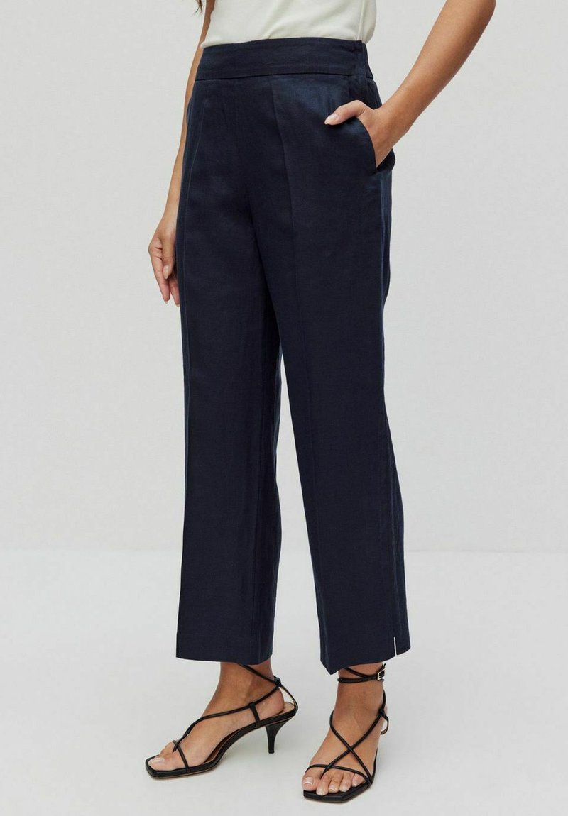 Granatowe lniane spodnie 7/8 z wysokim stanem, prostą nogawką i bocznymi kieszeniami. Charakteryzują się minimalnymi detalami szwów i sięgają nad kostkę.