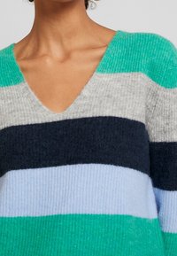 Pull-over tricoté avec un col en V, présentant des rayures horizontales en bleu sarcelle, bleu marine, bleu clair, gris et vert accentué. Tissu doux et texturé.