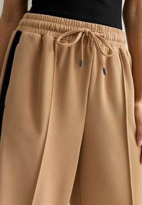 Pantaloni beige a pieghe con elastico in vita e cordino, caratterizzati da accenti laterali neri e una texture liscia. Estremità del cordino con punta in argento.