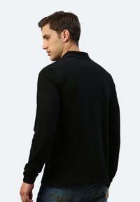 Schwarzes Langarm-Polohemd mit Kragen, strukturiertem Stoff und subtilen Nähdetails. Getragen mit blauen Jeans.