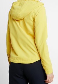 Gul letvægtsjakke med hætte, der har en glat tekstur, elastiske manchetter og en lige søm. Rygvisningen viser et enkelt design.