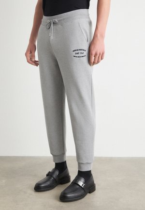 Person trägt hellgraue Sweatpants mit Seitentaschen und schwarzem Textlogo am Oberschenkel, kombiniert mit schwarzen Socken und schwarzen Leder-Loafers.