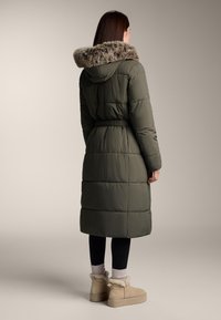 Dlhý olivovo-zelený páperový kabát s kapucňou podšitým kožušinou, s opaskom v páse, s prešívaným dizajnom, skombinovaný s svetlými hnedými čižmami s kožušinkou.