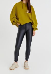 Sudadera oversized verde oliva con bolsillo tipo canguro, leggings negros de talle alto con acabado brillante y zapatillas color beige claro.