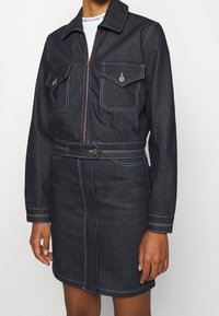 Denimjacka och kjol i mörk blå färg. Har två framfickor, dragkedjeslås och kontrasterande flerfärgade sömmar.