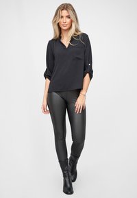Schwarze Bluse mit V-Ausschnitt und Tasche, kombiniert mit engen schwarzen Leggings. Das Modell trägt schwarze Stiefeletten. Glatte Textur, lässiges Design.