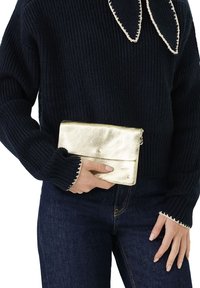 Henkilö pakahtaa pienen metallinhohtoisen kultapussin, jossa on tähden emblemia, ja hänellä on yllään tummansininen neulepaita valkoisilla reunoilla sekä tummansiniset farkut.