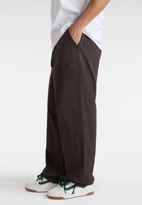 Brown wide-leg pants made of textured fabric, featuring side pockets and a small embroidered brand logo. Paired with white and green sneakers.