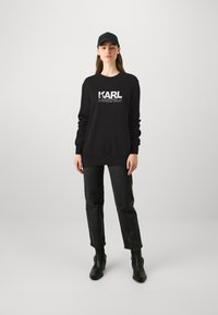 Čierna mikina s logom "KARL LAGERFELD" v bielej farbe, s voľným strihom, rebrovanými manžetami, skombinovaná s tmavými denimovými nohavicami a čiernymi čižmami.