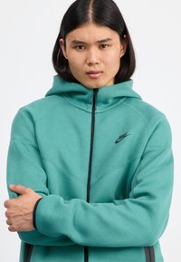 Mint green hoodie with a black zipper, Nike logo on the left chest, designed with a relaxed fit and ribbed cuffs, featuring soft fabric.