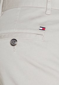 Hellgraue Hose mit glatter Textur, ausgestattet mit einem silbernen Knopf und einem kleinen gestickten rot, weiß und blau Logo in der Nähe der Tasche.