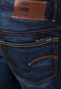 Mörkblå denimjeans med en läderlogotypmärke, gul söm och två bakfickor med märkesspecifika detaljer.