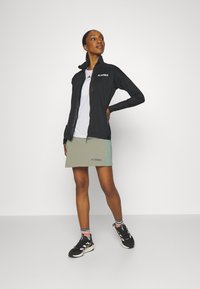 Schwarze Reißverschlussjacke, weißes Shirt und olivgrüne Skort mit blauem Logo. Schwarze Sportschuhe und mehrfarbige Socken runden das Outfit ab.