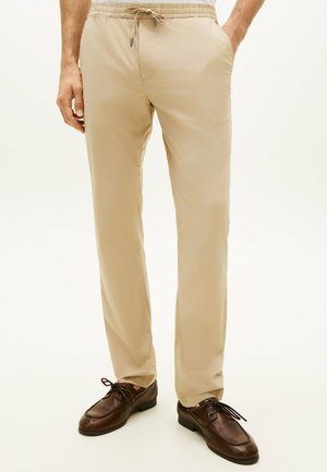 Mężczyzna w beżowych spodniach ze sznurkiem i ciemnobrązowych skórzanych butach sznurowanych, stojący z jedną ręką w kieszeni.