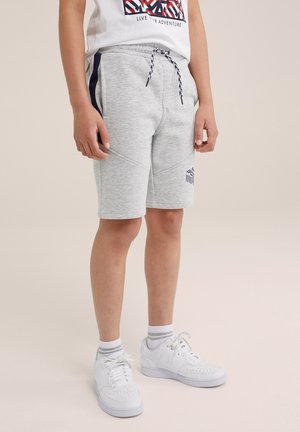 Kind trägt hellgraue knielange Shorts mit Kordelzug, weiße Turnschuhe und weiße Socken, steht vor einem schlichten Hintergrund.